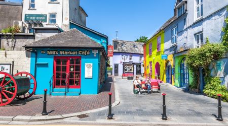 Kleurrijke straatscène die doet denken aan reizen Irland, met levendige gebouwen in tinten blauw, paars, geel en groen. Het "Milk Market Café" valt op als mensen op rode bankjes zitten. Een hoog grijs gebouw met het label "Grey Hound" doemt op in de achtergrond met een groot rood wiel aan de linkerkant onder een zonnige hemel. | Echt Ierland Vakanties