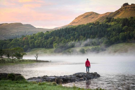 Een persoon in een rood jasje staat op een smal, rotsachtig pad dat uitloopt in een kalm meer tijdens hun rondreis Ierland. Mist hangt boven het water, met op de achtergrond weelderige groene heuvels en bergen onder een pastelroze en oranje zonsopganghemel. Bomen staan langs de overkant van dit serene Ierse landschap. | Echt Ierland Vakanties