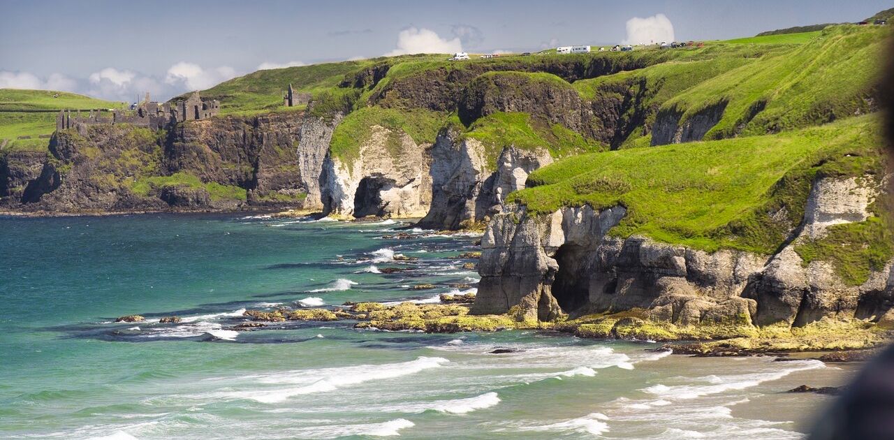 Ervaar het schilderachtige kustlandschap van een vakantie Ierland, met ruige kliffen versierd met groen gras, uitkijkend over een blauwgroene oceaan. Oude stenen ruïnes en een kronkelende weg verschijnen boven op de kliffen onder gedeeltelijk bewolkte luchten, perfect voor een onvergetelijke rondreis Ierland. | Echt Ierland Vakanties