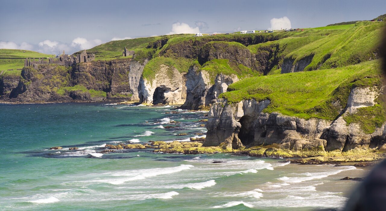 Ervaar het schilderachtige kustlandschap van een vakantie Ierland, met ruige kliffen versierd met groen gras, uitkijkend over een blauwgroene oceaan. Oude stenen ruïnes en een kronkelende weg verschijnen boven op de kliffen onder gedeeltelijk bewolkte luchten, perfect voor een onvergetelijke rondreis Ierland. | Echt Ierland Vakanties