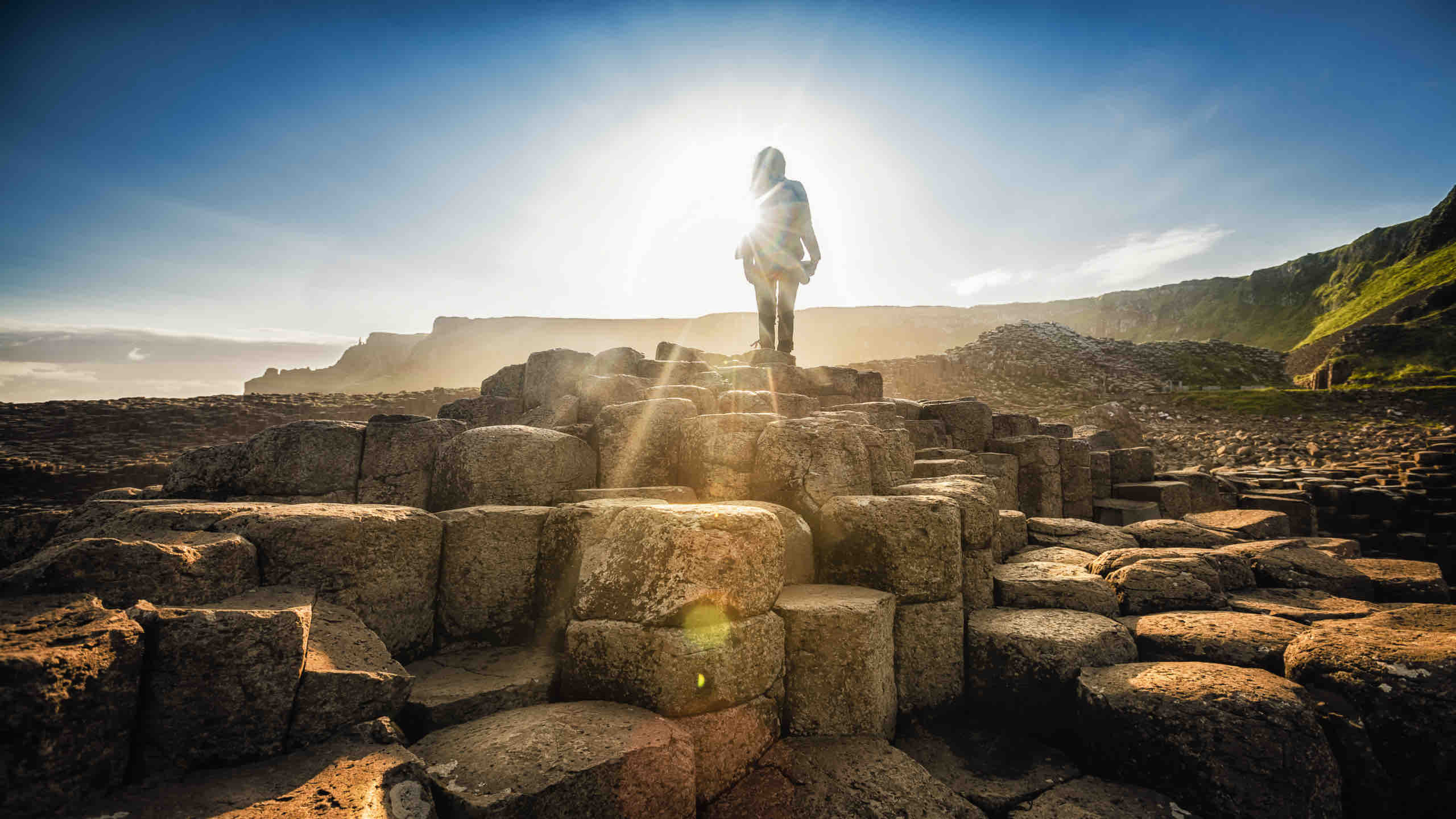 Een persoon staat bovenop de Giant’s Causeway in Noord-Ierland, met de zon die een dramatisch silhouet werpt. Omringd door hexagonale basaltkolommen onder een helderblauwe lucht, is dit magische moment slechts een hoogtepunt van een onvergetelijk rondreis Ierland-avontuur. | Echt Ierland Vakanties