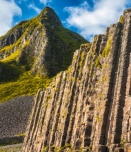Een ruig landschap met hoge, hexagonale basaltkolommen aan de rechterkant, mogelijk gevormd door vulkanische activiteit. Dit adembenemende tafereel is typerend voor een rondreis Ierland, waar de kolommen steil oprijzen tegen weelderige groene heuvels en grillige pieken onder een blauwe lucht met verspreide witte wolken. | Echt Ierland Vakanties
