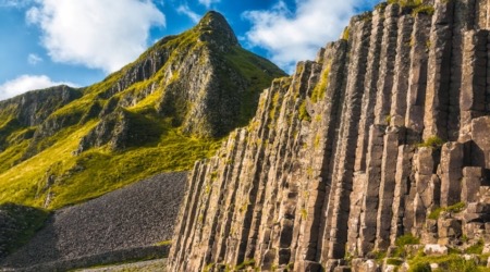 Een ruig landschap met hoge, hexagonale basaltkolommen aan de rechterkant, mogelijk gevormd door vulkanische activiteit. Dit adembenemende tafereel is typerend voor een rondreis Ierland, waar de kolommen steil oprijzen tegen weelderige groene heuvels en grillige pieken onder een blauwe lucht met verspreide witte wolken. | Echt Ierland Vakanties