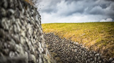 Een stenen muur met een smalle doorgang omlijnt een met gras begroeide heuvel, en legt de ruige schoonheid van een Iers landschap vast. De ruwe textuur van de stenen contrasteert met het gladde, groenachtig-gele gras, terwijl grijze wolken wijzen op dreigende regen - een serene uitnodiging om Ierland te verkennen tijdens uw vakantie. | Echt Ierland Vakanties