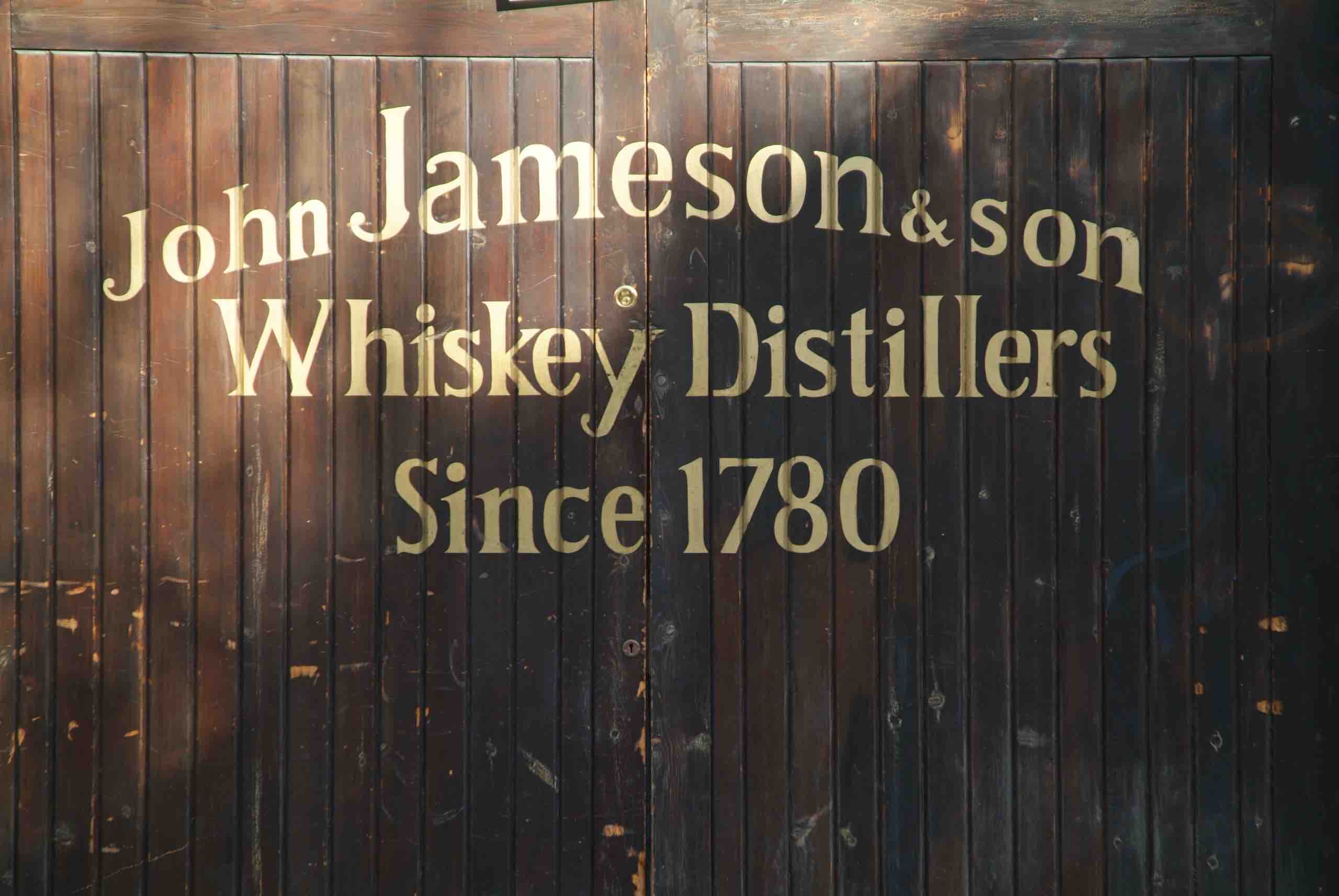 Donkere houten deur met gouden tekst "John Jameson & Son Whiskey Distillers Since 1780." De oude en versleten look voegt rustieke charme toe, perfect voor degenen die op een rondreis Ierland op zoek zijn naar historische en traditionele ervaringen. De gecentreerde, licht gebogen tekst versterkt de tijdloze aantrekkingskracht. | Echt Ierland Vakanties
