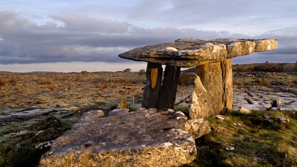 Oud stenen portaalgraf met grote horizontale platen op verticale steunen, genesteld in een grasachtig, rotsachtig Iers landschap onder een bewolkte zonsondergang. De verweerde stenen versterken het historische gevoel - een must-see op elke vakantie in Ierland voor degenen die gefascineerd zijn door tijdloze schoonheid. | Echt Ierland Vakanties