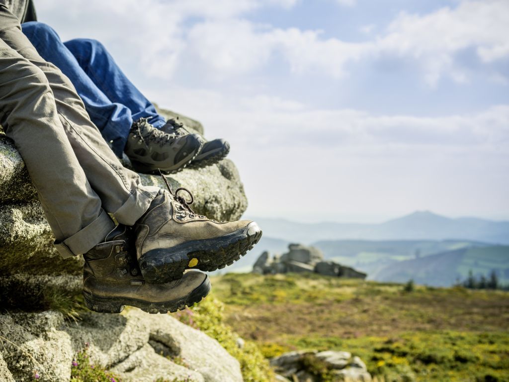 Close-up van twee paar benen die aan een rotsachtige richel hangen tijdens een vakantie in Ierland. Op de voorgrond zijn bruine wandelschoenen te zien, terwijl op de achtergrond zwarte en groene schoenen te zien zijn. Een schilderachtig landschap met glooiende heuvels, groene velden en een bewolkte lucht strekt zich daarachter uit. | Echt Ierland Vakanties