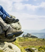 Close-up van twee paar benen die aan een rotsachtige richel hangen tijdens een vakantie in Ierland. Op de voorgrond zijn bruine wandelschoenen te zien, terwijl op de achtergrond zwarte en groene schoenen te zien zijn. Een schilderachtig landschap met glooiende heuvels, groene velden en een bewolkte lucht strekt zich daarachter uit. | Echt Ierland Vakanties
