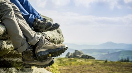 Close-up van twee paar benen die aan een rotsachtige richel hangen tijdens een vakantie in Ierland. Op de voorgrond zijn bruine wandelschoenen te zien, terwijl op de achtergrond zwarte en groene schoenen te zien zijn. Een schilderachtig landschap met glooiende heuvels, groene velden en een bewolkte lucht strekt zich daarachter uit. | Echt Ierland Vakanties