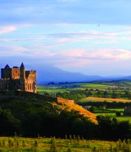 Een schilderachtig uitzicht op de Rock of Cashel, een historisch kasteel op een heuvel, verlicht door de ondergaande zon tijdens uw rondreis Ierland. Weelderige groene velden en verre glooiende heuvels strekken zich uit onder een gedeeltelijk bewolkte hemel. Het rustige landschap benadrukt de grandeur van het oude bouwwerk. | Echt Ierland Vakanties