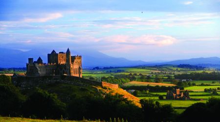 Een schilderachtig uitzicht op de Rock of Cashel, een historisch kasteel op een heuvel, verlicht door de ondergaande zon tijdens uw rondreis Ierland. Weelderige groene velden en verre glooiende heuvels strekken zich uit onder een gedeeltelijk bewolkte hemel. Het rustige landschap benadrukt de grandeur van het oude bouwwerk. | Echt Ierland Vakanties