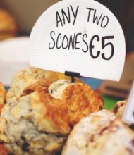 Een selectie scones op een bruisende Ierse markt verleidt reizigers. Op een bord staat "Twee willekeurige scones €5." Kleinere borden pochen "Giant blueberry scones with fresh blueberries €3." Het assortiment, rijk aan texturen en kleuren, is een uitnodigende stop op elk rondreis Ierland avontuur. | Echt Ierland Vakanties