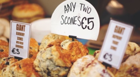 Een selectie scones op een bruisende Ierse markt verleidt reizigers. Op een bord staat "Twee willekeurige scones €5." Kleinere borden pochen "Giant blueberry scones with fresh blueberries €3." Het assortiment, rijk aan texturen en kleuren, is een uitnodigende stop op elk rondreis Ierland avontuur. | Echt Ierland Vakanties