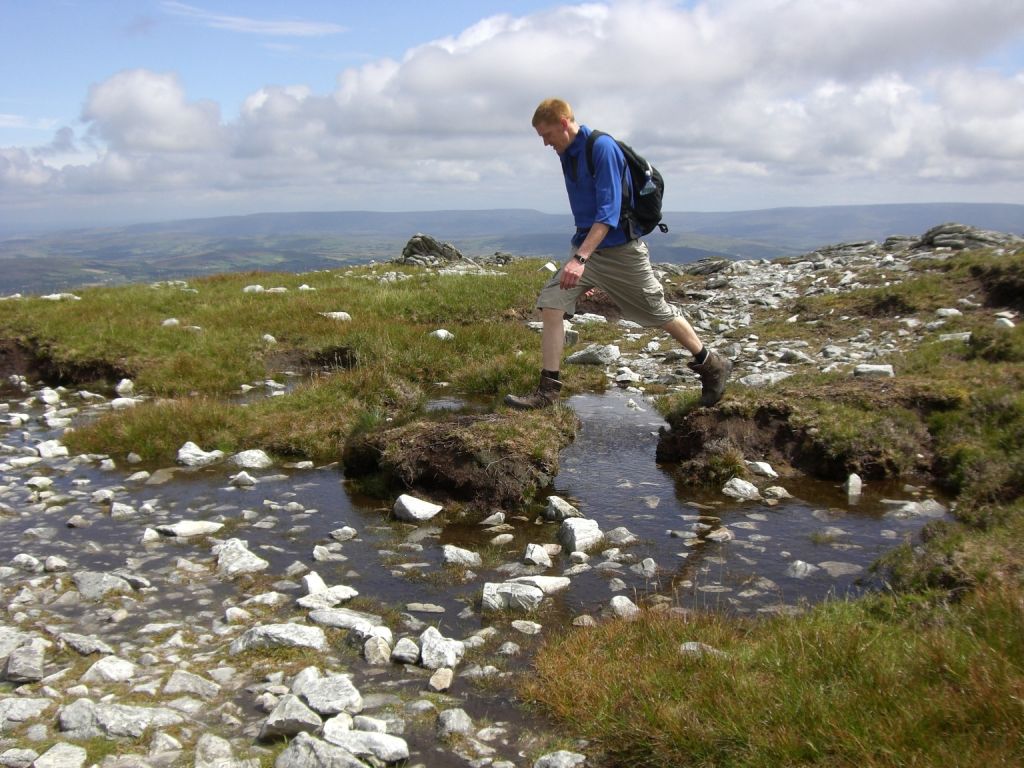 Een persoon met kort haar wandelt over een rotsachtig pad in een grasachtig, bergachtig gebied op hun vakantie in Ierland. Ze dragen een blauw shirt met lange mouwen, een kaki broek en wandelschoenen, met een zwarte rugzak. Ze stappen over een ondiepe stroom tussen grasvelden en verspreide witte rotsen onder een Ierse bewolkte lucht. | Echt Ierland Vakanties