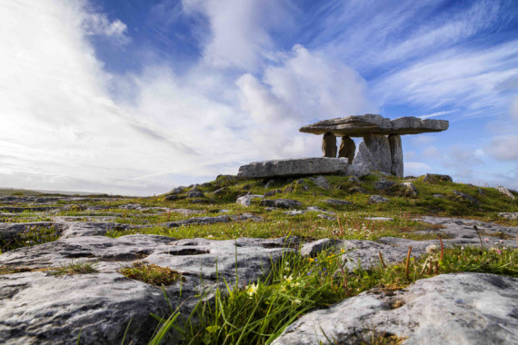 Onder een helderblauwe lucht met verspreide wolken staat een eeuwenoude stenen dolmen als een getuigenis van de geschiedenis, perfect voor degenen die een rondreis door Ierland maken. De voorgrond toont ruige rotsen en stukken groen gras bezaaid met kleine gele bloemen, die dit prehistorische monument aanvullen. | Echt Ierland Vakanties
