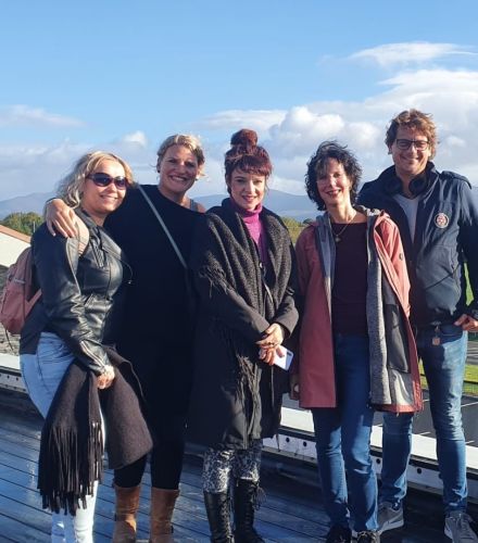 Vijf mensen staan dicht bij elkaar op een houten platform buiten tegen een achtergrond van verre bergen en een gedeeltelijk bewolkte lucht, en vangen de vreugde van hun rundreise Ierland. Ze zijn gekleed in casual, warme kleding, inclusief jassen en sjaals, allemaal lachend en naast elkaar staand. | Echt Ierland Vakanties