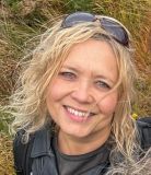 Een vrouw met krullend blond haar en blauwe ogen lacht naar de camera. Ze draagt een zonnebril op haar hoofd en een zwart jasje, wat suggereert dat ze geniet van een rondreis Ierland. De achtergrond van hoog gras en groen vangt de charme van een Ierse buitenomgeving. | Echt Ierland Vakanties