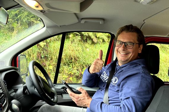 Een glimlachende persoon met een bril en een blauwe hoodie zit op de bestuurdersstoel van een rood voertuig en steekt zijn duim omhoog. Ze houden een smartphone vast, misschien plannen ze hun volgende urlaub Irland-avontuur. Het raam achter hen laat weelderig groen gebladerte en planten zien. | Echt Ierland Vakanties