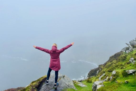 Een persoon in een kastanjebruin jasje staat op een rotsachtige, met gras begroeide klif met uitgestrekte armen, uitkijkend over het mistige landschap van een rundreise Ierland. De oceaan eronder is nauwelijks zichtbaar door de dichte mist, wat het gevoel van uitgestrektheid en isolatie versterkt - een ware belichaming van vrijheid en verkenning. | Echt Ierland Vakanties