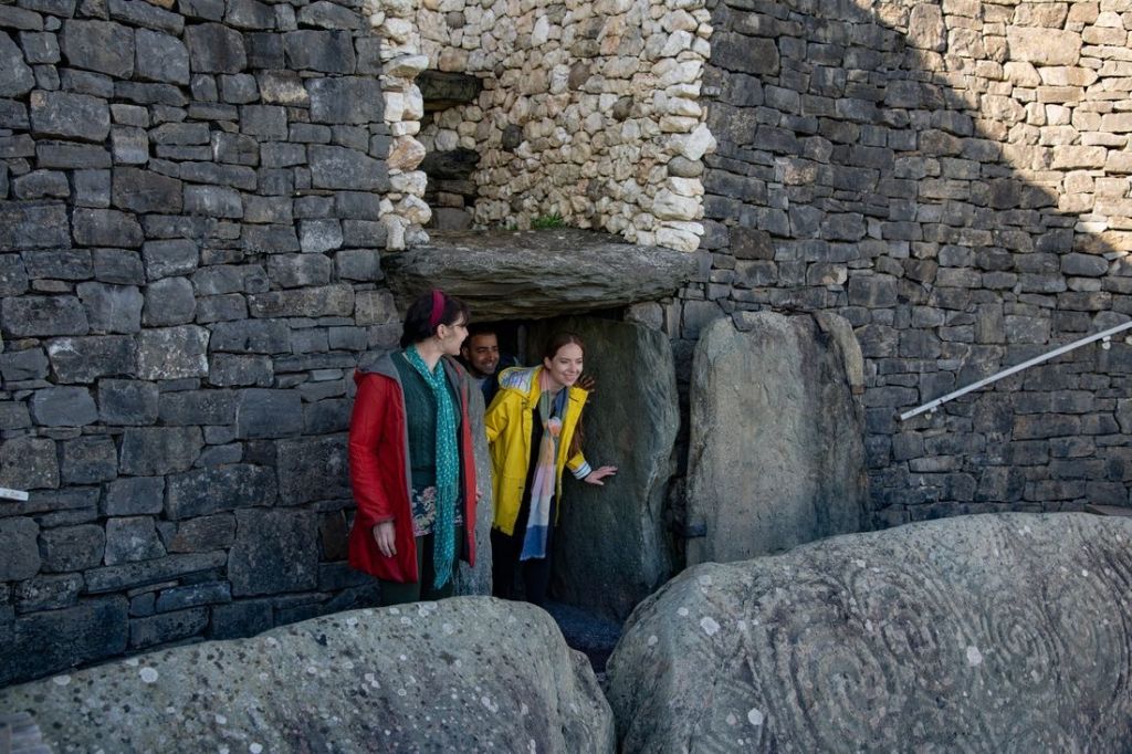 Drie mensen komen uit een oude stenen doorgang op hun rundreise Irland. De muren, gemaakt van grote, ronde en plaatachtige stenen, vormen een opvallende achtergrond tegen hun kleurrijke jassen. Op de voorgrond ligt een gegraveerde steen met cirkelvormige patronen, verlicht door natuurlijk licht van buitenaf. | Echt Ierland Vakanties