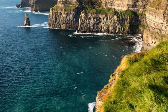 Ervaar het adembenemende uitzicht op de Cliffs of Moher tijdens uw vakantie in Ierland. Steile rotsachtige kliffen rijzen majestueus uit boven de oceaan, met weelderig groen gras op de voorgrond en een kalme zee onder een gedeeltelijk bewolkte lucht. Een geïsoleerde rotsformatie staat trots bij de kliffen. | Echt Ierland Vakanties