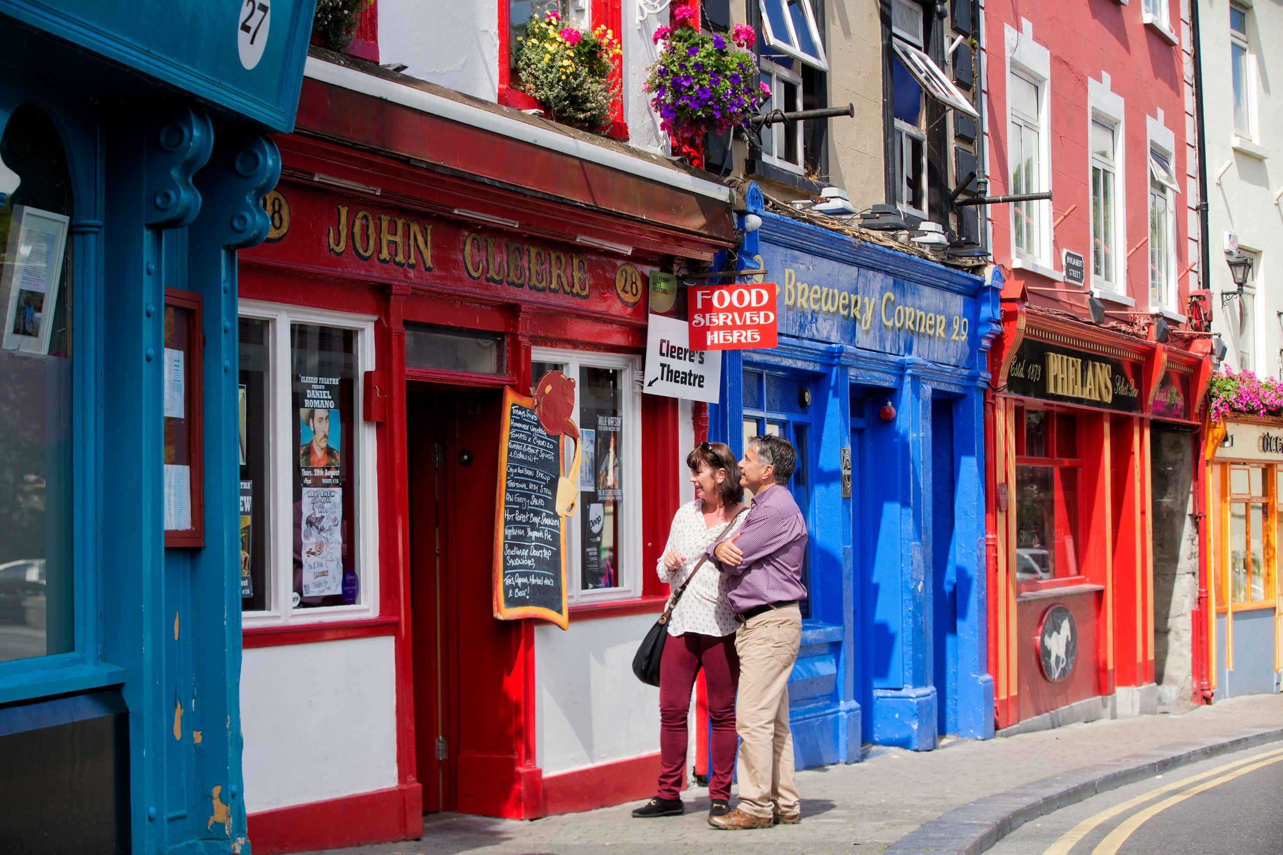 Een stel staat buiten een kleurrijke straat met levendige winkels en restaurants, die de charme van een schilderachtige Europese sfeer belichamen. Ze bestuderen een menu op een rode pub met gele bewegwijzering, die doet denken aan veel op een rundreise Ierland, terwijl blauwe en bruine gebouwen pronken met vensterbanken vol bloemen. | Echt Ierland Vakanties