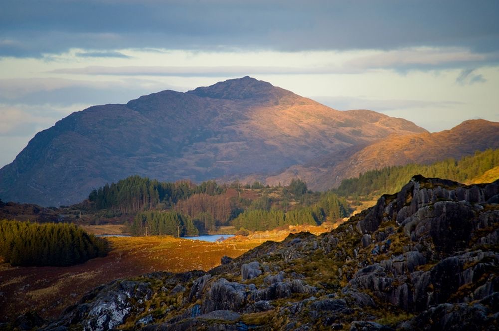 Een berglandschap met zacht zonlicht dat over het rotsachtige terrein schijnt, lijkt op de serene schoonheid die je zou kunnen vinden tijdens een reis naar Ierland. Een weelderig bos aan de voet van de berg en een vijver die de lucht weerspiegelt onder gedeeltelijk bewolkte plekken creëren een idyllisch tafereel verlicht door gouden zonlicht. | Echt Ierland Vakanties