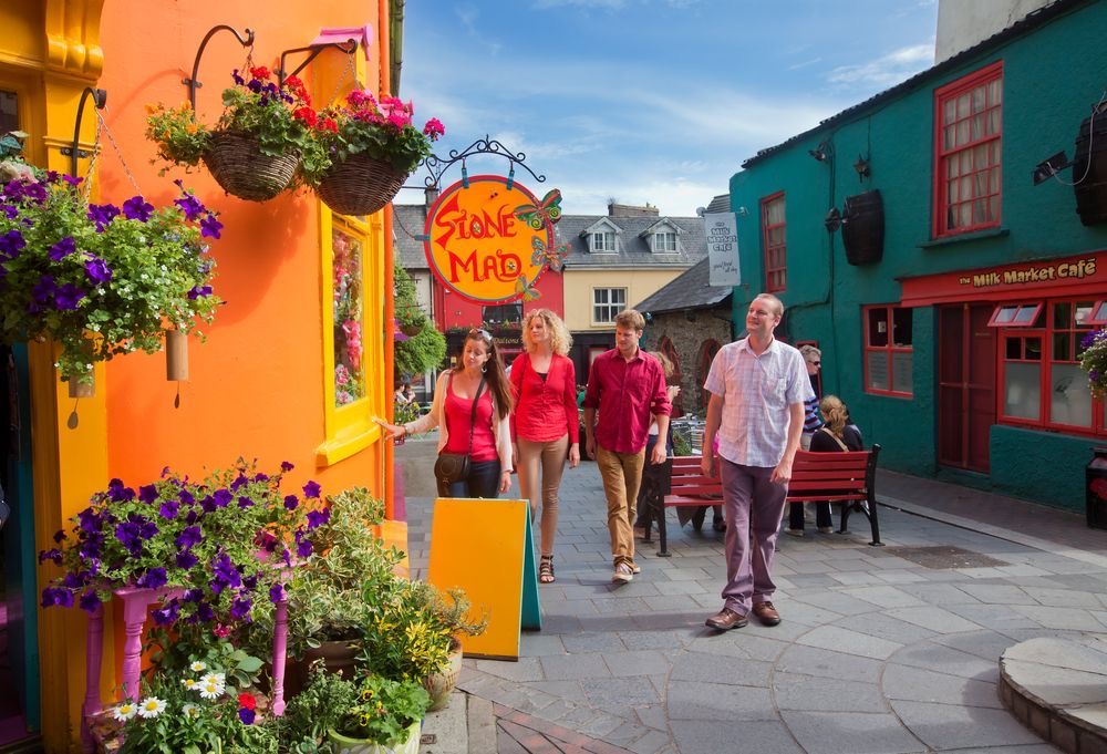 Een groep van vier mensen wandelt door een levendige straat tijdens hun vakantie in Ierland. De kleurrijke gebouwen omvatten een oranje met een "Stone Mad"-bord en hangende bloemenmanden. Aqua en rode tinten sieren andere, terwijl een café zichtbaar is. De lucht is blauw, met wolken boven het geplaveide pad omzoomd door plantenbakken. | Echt Ierland Vakanties