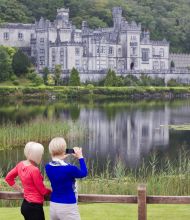 Twee mensen staan bij een houten hek, degene links in een rode trui en degene rechts in een blauwe sjaal met een camera in de hand. Ze bewonderen een groot, rijk versierd kasteel aan de overkant van een sereen meer, dat hun urlaub Ierland belichaamt te midden van weelderig groen met reflecties die dansen in het water. | Echt Ierland Vakanties