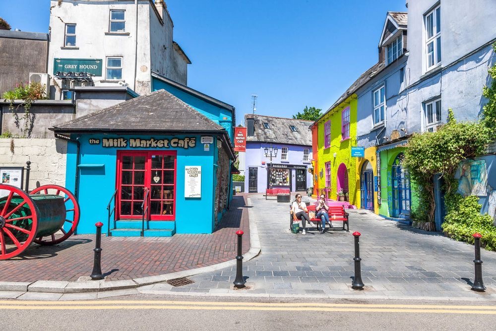 Een levendig straatbeeld in Ierland met kleurrijke gebouwen. Het Milk Market Café, turquoise geverfd, staat prominent aan de linkerkant. Felgele en blauwe gebouwen staan aan de rechterkant. Mensen ontspannen op bankjes onder een heldere hemel, terwijl een vintage rode kar charme toevoegt - een perfect momentopname van uw vakantie in Ierland. | Echt Ierland Vakanties