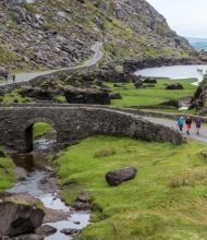 Een kronkelende weg en stenen brug doorkruisen een weelderig groen landschap met rotsachtige heuvels in dit idyllische tafereel, perfect voor *Urlaub Irland*. Een smalle beek stroomt onder de brug door. Drie mensen lopen langs de weg. Twee kleine meren zijn zichtbaar, omringd door gras en keien onder een bewolkte lucht. | Echt Ierland Vakanties