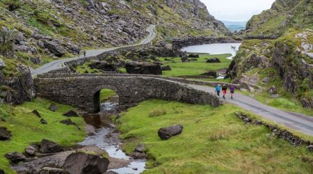 Een kronkelende weg en stenen brug doorkruisen een weelderig groen landschap met rotsachtige heuvels in dit idyllische tafereel, perfect voor *Urlaub Irland*. Een smalle beek stroomt onder de brug door. Drie mensen lopen langs de weg. Twee kleine meren zijn zichtbaar, omringd door gras en keien onder een bewolkte lucht. | Echt Ierland Vakanties