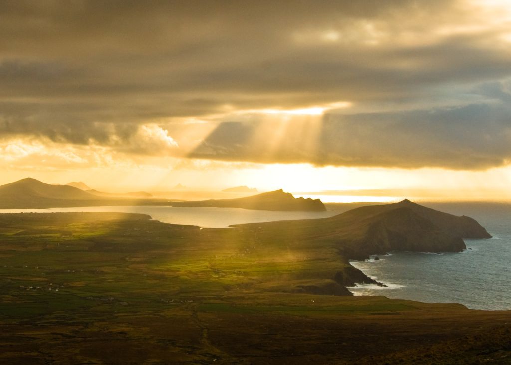Zonnelicht breekt door dramatische wolken boven de ruige kustlijn van een schiereiland en werpt gouden stralen op de groene velden van Ierland eronder. Perfect voor een rundreise Ierland, de oceaan weerkaatst het licht, terwijl verre heuvels en landvormen de horizon onder een bewolkte lucht omlijnen. | Echt Ierland Vakanties