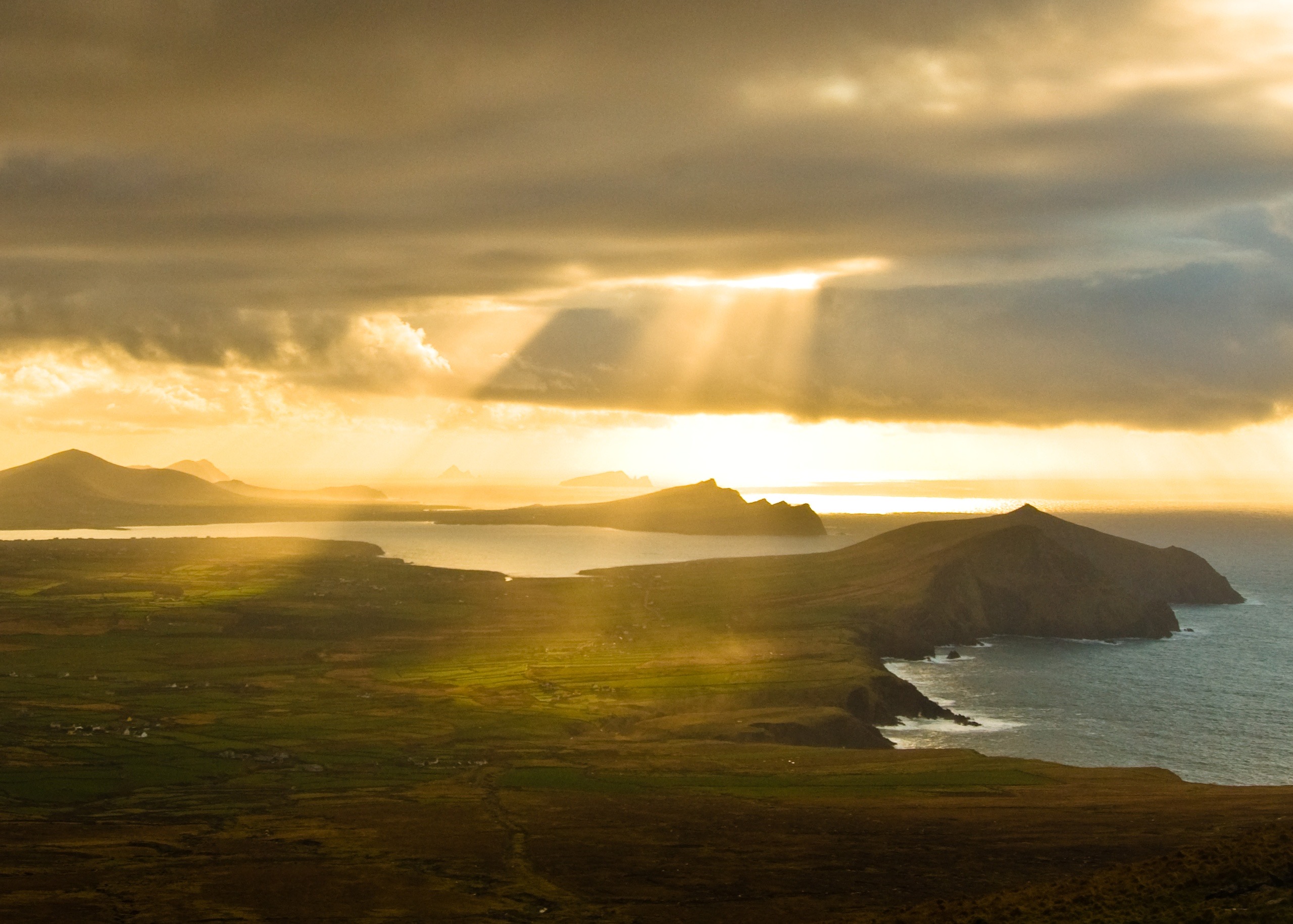 Zonnelicht breekt door dramatische wolken boven de ruige kustlijn van een schiereiland en werpt gouden stralen op de groene velden van Ierland eronder. Perfect voor een rundreise Ierland, de oceaan weerkaatst het licht, terwijl verre heuvels en landvormen de horizon onder een bewolkte lucht omlijnen. | Echt Ierland Vakanties
