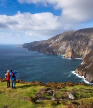 Twee wandelaars staan op een met gras begroeide klifrand met uitzicht op de oceaan bij Slieve League in Ierland. Een wijst naar torenhoge kliffen die de zee raken onder een gedeeltelijk bewolkte lucht. Perfect voor elke rondreis Ierland, het landschap bestaat uit ruig terrein met stukken gras en struikgewas. | Echt Ierland Vakanties