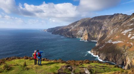 Twee wandelaars staan op een met gras begroeide klifrand met uitzicht op de oceaan bij Slieve League in Ierland. Een wijst naar torenhoge kliffen die de zee raken onder een gedeeltelijk bewolkte lucht. Perfect voor elke rondreis Ierland, het landschap bestaat uit ruig terrein met stukken gras en struikgewas. | Echt Ierland Vakanties