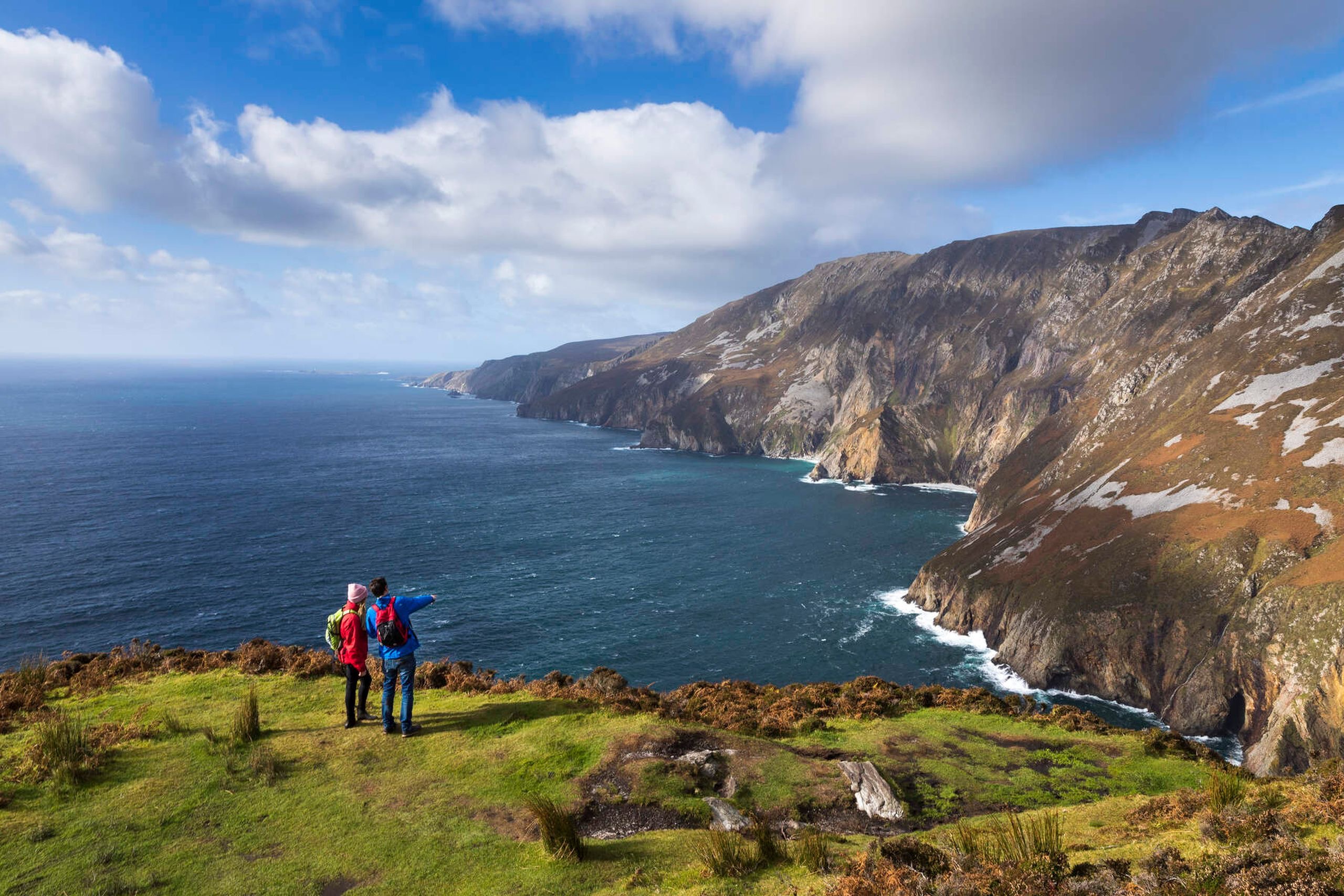 Twee wandelaars staan op een met gras begroeide klifrand met uitzicht op de oceaan bij Slieve League in Ierland. Een wijst naar torenhoge kliffen die de zee raken onder een gedeeltelijk bewolkte lucht. Perfect voor elke rondreis Ierland, het landschap bestaat uit ruig terrein met stukken gras en struikgewas. | Echt Ierland Vakanties