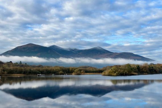 Een rustig meer in Ierland weerspiegelt een bewolkte lucht en verre bergen. Lage mist drijft boven het water en verbergt gedeeltelijk de beboste oever. Verschillende kleine boten liggen afgemeerd op het meer, wat dit vredige tafereel versterkt - een perfect hoogtepunt voor elke rundreise Ierland. | Echt Ierland Vakanties