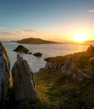 Een schilderachtig kustlandschap bij zonsondergang, dat doet denken aan de Rundreise in Ierland, met rotsachtige kliffen en grillige rotsen op de voorgrond. De zon gaat onder aan de horizon en werpt een gouden gloed over het kalme oceaanwater. De kleurovergang van oranje en blauw aan de hemel, met verspreide wolken, versterkt dit serene tafereel. | Echt Ierland Vakanties