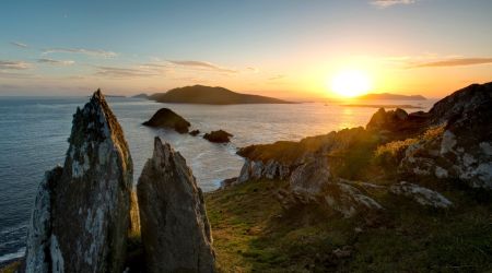 Een schilderachtig kustlandschap bij zonsondergang, dat doet denken aan de Rundreise in Ierland, met rotsachtige kliffen en grillige rotsen op de voorgrond. De zon gaat onder aan de horizon en werpt een gouden gloed over het kalme oceaanwater. De kleurovergang van oranje en blauw aan de hemel, met verspreide wolken, versterkt dit serene tafereel. | Echt Ierland Vakanties