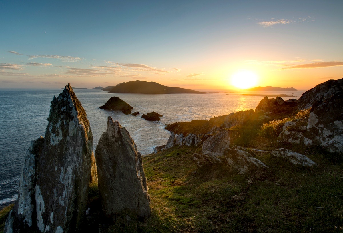 Een schilderachtig kustlandschap bij zonsondergang, dat doet denken aan de Rundreise in Ierland, met rotsachtige kliffen en grillige rotsen op de voorgrond. De zon gaat onder aan de horizon en werpt een gouden gloed over het kalme oceaanwater. De kleurovergang van oranje en blauw aan de hemel, met verspreide wolken, versterkt dit serene tafereel. | Echt Ierland Vakanties