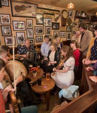 Een levendige groep muzikanten en toeschouwers in een gezellige pub, perfect voor degenen die op vakantie zijn in Ierland. Muzikanten bespelen traditionele Ierse instrumenten: viool, accordeon en drum. De muren zijn versierd met ingelijste foto's en kleine tafels met drankjes terwijl een divers publiek geniet van de muziek. | Echt Ierland Vakanties