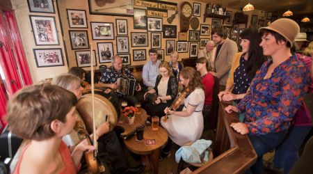 Een levendige groep muzikanten en toeschouwers in een gezellige pub, perfect voor degenen die op vakantie zijn in Ierland. Muzikanten bespelen traditionele Ierse instrumenten: viool, accordeon en drum. De muren zijn versierd met ingelijste foto's en kleine tafels met drankjes terwijl een divers publiek geniet van de muziek. | Echt Ierland Vakanties