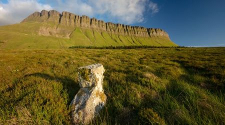 Een eenzame rots staat op een grasachtige voorgrond met wilde bloemen. Daarachter strekt een breed veld zich uit tot aan de ruige bergketen onder een helderblauwe lucht, die doet denken aan een betoverende urlaub Ierland. De kliffen van de bergen zijn opvallend en de verlichting suggereert vroege ochtend of late middag. | Echt Ierland Vakanties