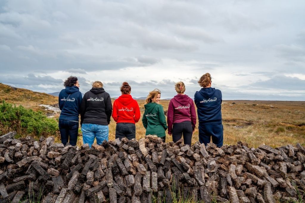 Zes mensen staan met hun rug naar de camera, kijkend naar een uitgestrekt, bewolkt landschap met gras en rotsen tijdens hun rondreis Ierland. Ze dragen kleurrijke hoodies met tekst op de achterkant en zitten op een stapel turf. De bewolkte lucht vormt een dramatische achtergrond voor de aardse tinten van dit Ierse tafereel. | Echt Ierland Vakanties