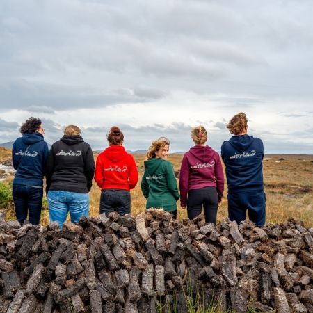 Zes mensen staan met hun rug naar elkaar toe, met hoodies in zwart, rood, groen en marineblauw. Ze kijken uit over een uitgestrekt graslandschap onder een bewolkte lucht, wat de geest van een authentieke vakantie in Ierland belichaamt. Voor hen ligt een stapel gekapte turfstenen. | Echt Ierland Vakanties