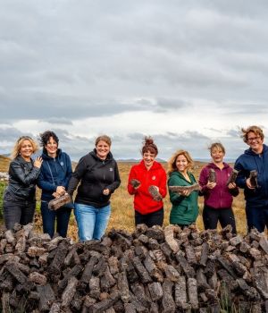 Zeven mensen staan achter een grote stapel turfstenen in een graslandschap onder een bewolkte lucht. Lachend en gekleed in buitenkleding, houdt ieder een stuk turf vast, wat de geest van gemeenschappelijke activiteit belichaamt. Deze scène legt de essentie van een authentieke reisen Irland-ervaring vast. | Echt Ierland Vakanties