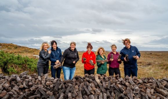 Zeven mensen staan achter een grote stapel turfstenen in een graslandschap onder een bewolkte lucht. Lachend en gekleed in buitenkleding, houdt ieder een stuk turf vast, wat de geest van gemeenschappelijke activiteit belichaamt. Deze scène legt de essentie van een authentieke reisen Irland-ervaring vast. | Echt Ierland Vakanties