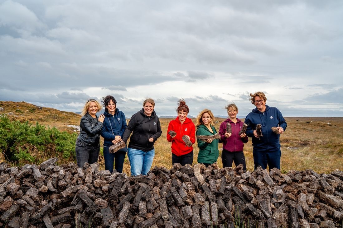Zeven mensen staan achter een grote stapel turfstenen in een graslandschap onder een bewolkte lucht. Lachend en gekleed in buitenkleding, houdt ieder een stuk turf vast, wat de geest van gemeenschappelijke activiteit belichaamt. Deze scène legt de essentie van een authentieke reisen Irland-ervaring vast. | Echt Ierland Vakanties