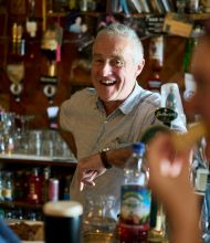 Een oudere man met een vrolijke uitdrukking staat zelfverzekerd achter een bar, die doet denken aan een gezellige pub op een rondreis Ierland. Omringd door flessen en barparafernalia weerspiegelt de warme, gezellige setting de uitnodigende charme van een onvergetelijke vakantie Ierland. | Echt Ierland Vakanties